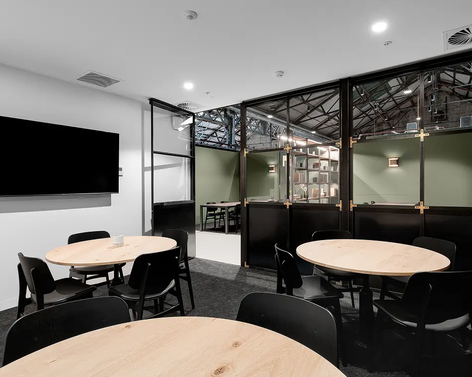 Light wood round tables surrounded by black chairs sit on a dark carpet inside a modern office room equipped with a large wall-mounted TV screen and glass partitions.
