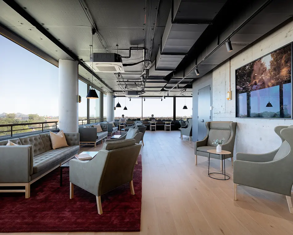 Modern grey sofas and wingback armchairs furnish a spacious, open-plan lounge with floor-to-ceiling windows, dark exposed ceiling ducts, light wood flooring, and a burgundy area rug.
