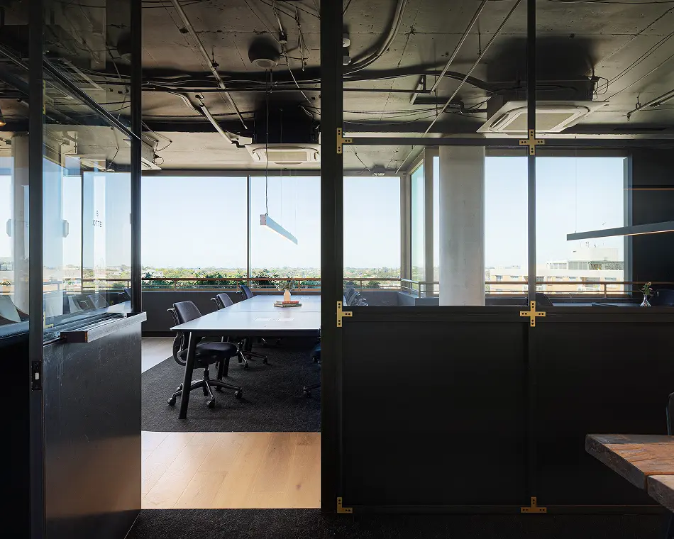 A long conference table surrounded by black rolling chairs stands inside a glass-walled meeting room with exposed ceiling pipes and floor-to-ceiling windows overlooking a city view. Text on glass reads: "8 OTTE".