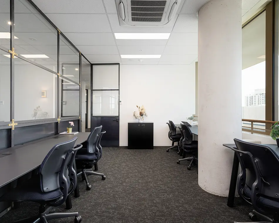 Black ergonomic office chairs sit at sleek dark desks arranged in a modern office space featuring dark carpeting, glass-paneled walls, a large concrete pillar, and bright ceiling lights.