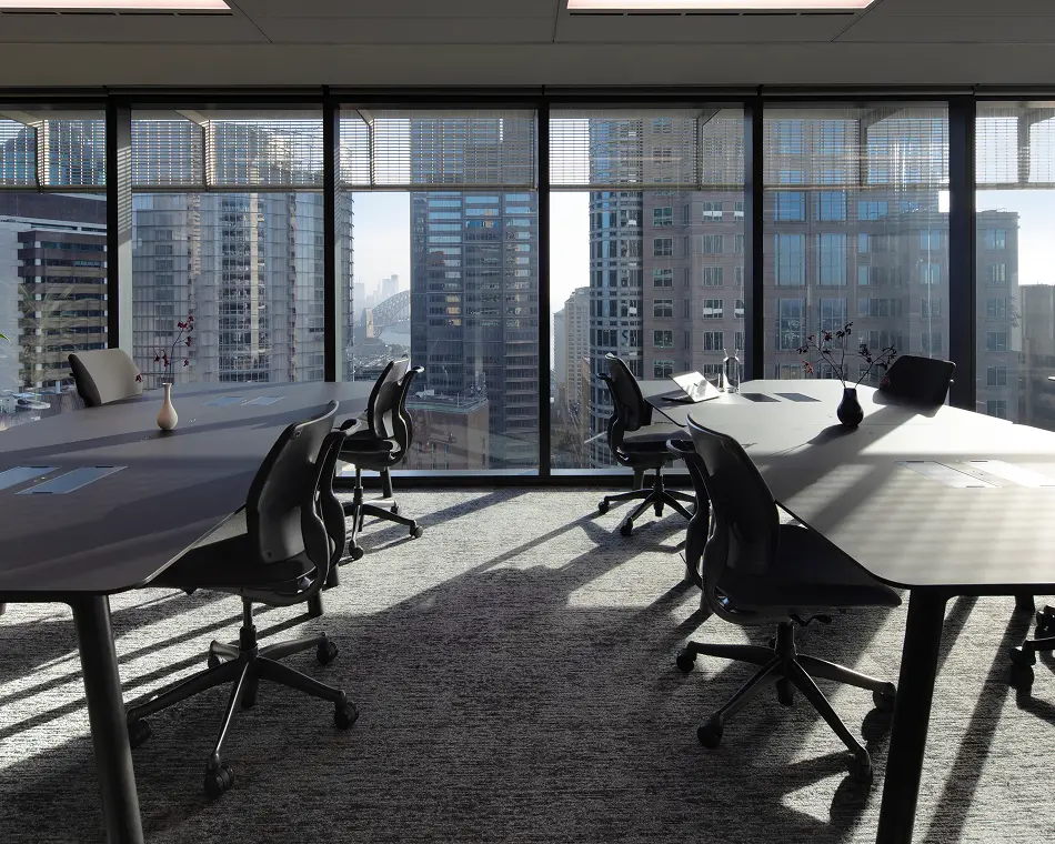 Sleek office desks surrounded by ergonomic chairs sit in a sunny high-rise room, casting sharp shadows across carpeted floors before floor-to-ceiling windows that overlook a dense skyscraper cityscape.