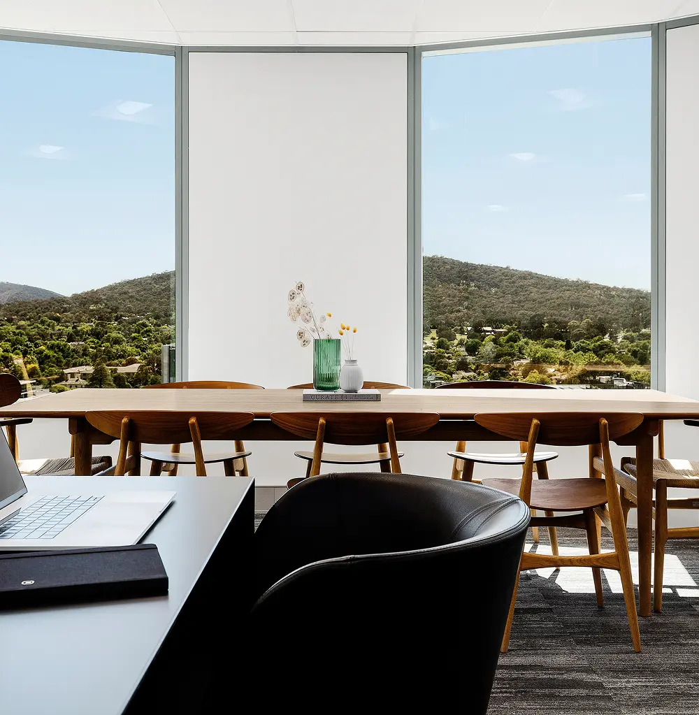 A long wooden conference table surrounded by chairs stands before floor-to-ceiling windows overlooking lush green hills. A book resting on the table displays the text "CURATE".