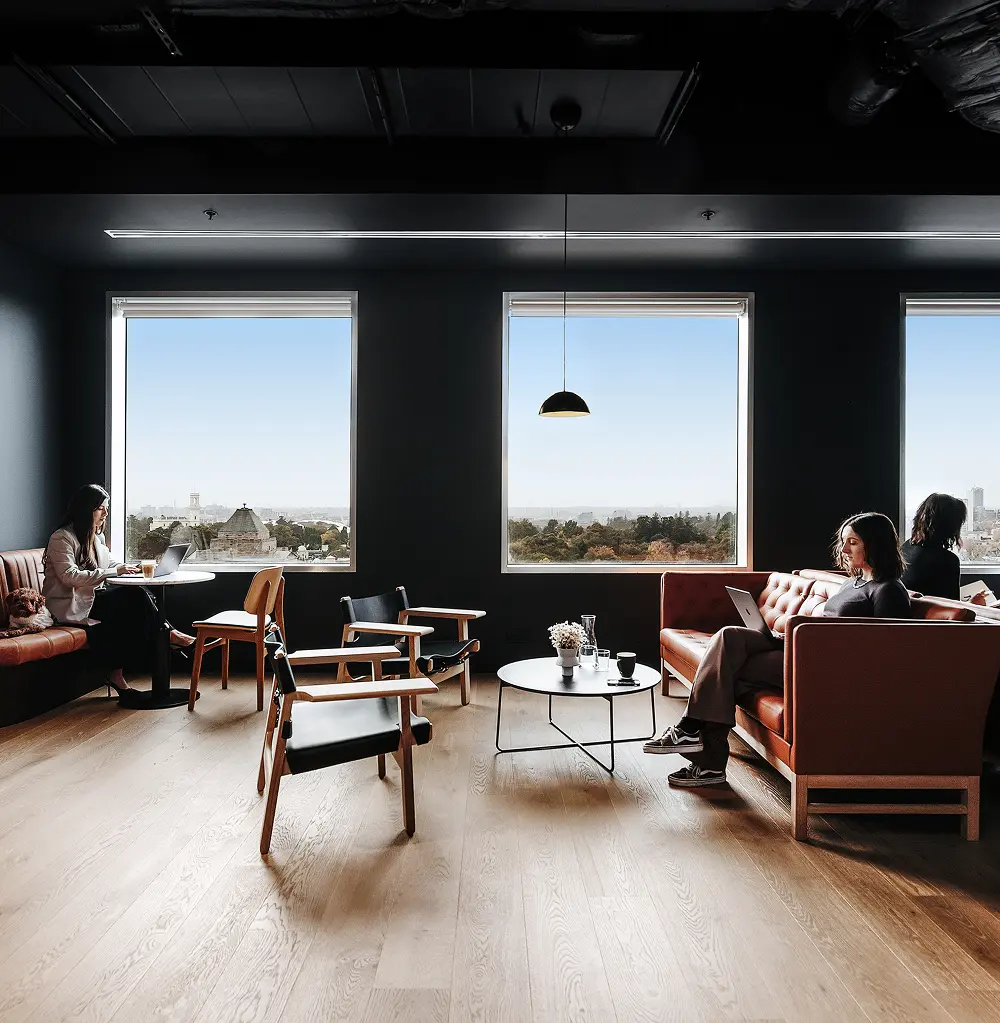 People sit on leather sofas and wooden armchairs working on laptops inside a dark, modern office lounge featuring large square windows that look out onto a city park.