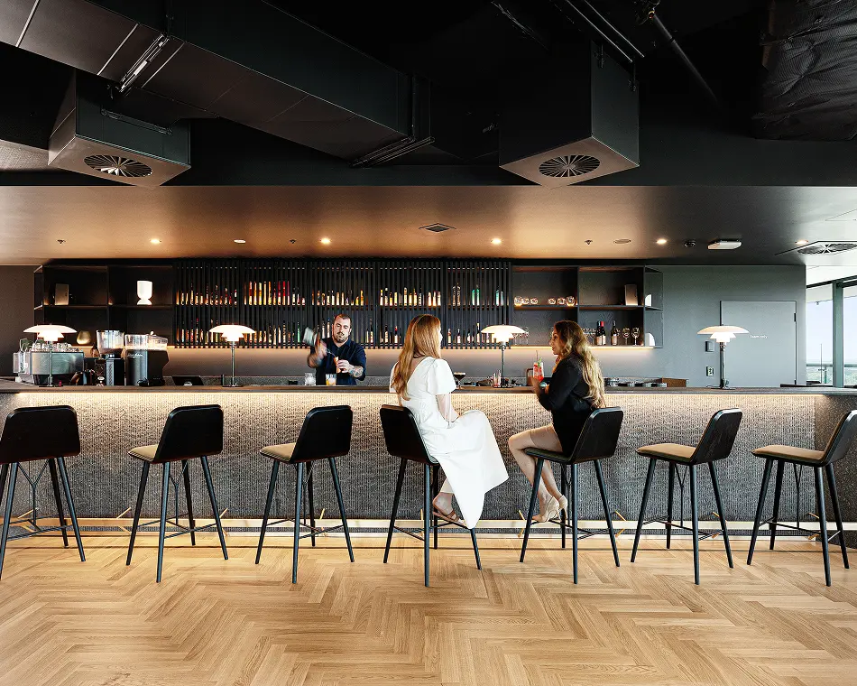 Two women sit on barstools and converse while a male bartender mixes a drink behind an illuminated counter within a modern lounge with herringbone wood floors. Text on the right door reads, “Team only”.