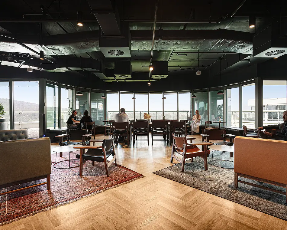 People sit, work, and converse inside a spacious modern lounge featuring herringbone wooden floors, stylish seating on patterned rugs, exposed industrial ceiling ducts, and expansive floor-to-ceiling windows.