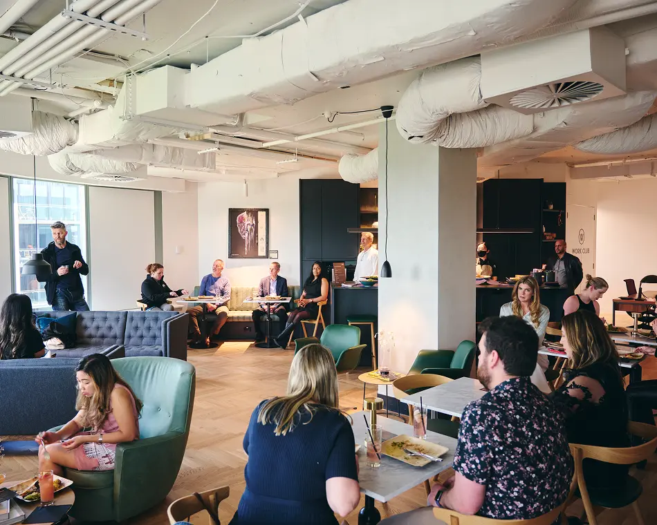 People sit and converse at tables and armchairs within a modern, industrial office lounge featuring exposed ceiling ductwork. Text on the right background wall reads, "WORK CLUB".