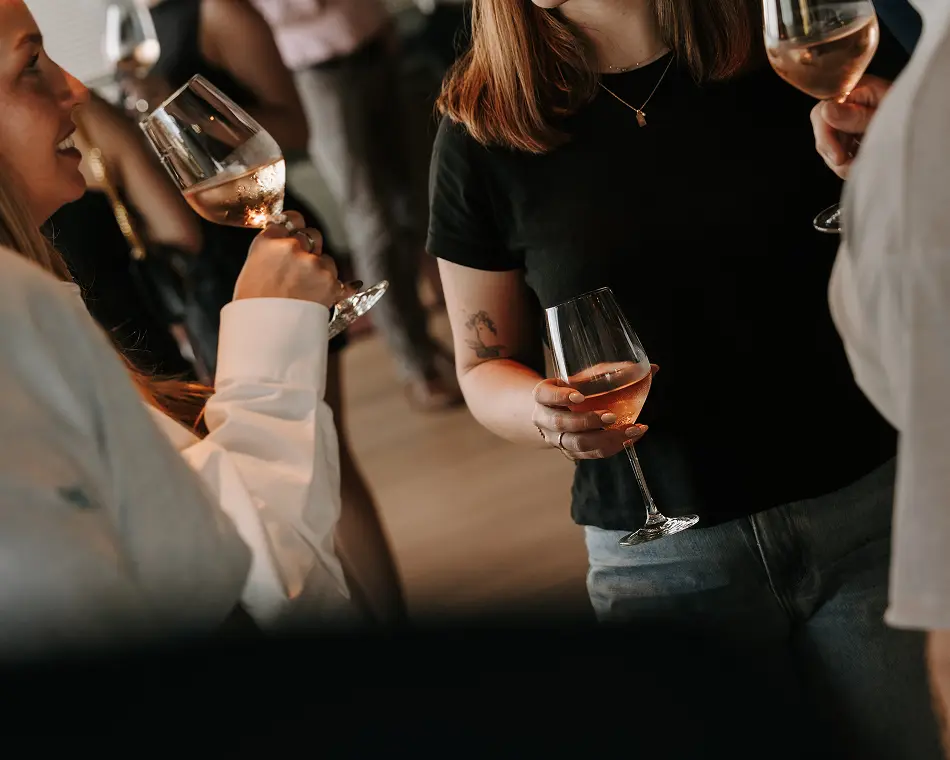 People hold and raise wine glasses filled with rosé while socializing and mingling together during an indoor party.