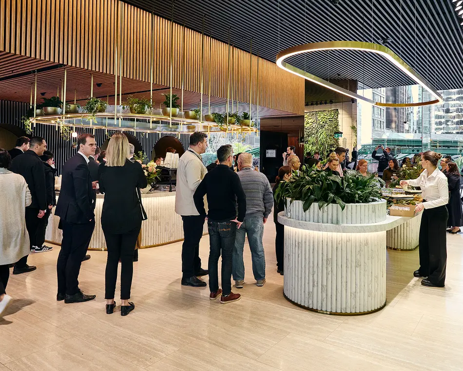 People in business attire mingle and gather around marble service counters inside a modern, upscale lobby featuring hanging brass planters and slatted wooden ceilings.