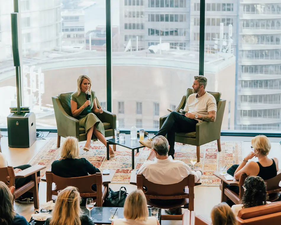 A woman speaking into a microphone sits alongside a man in armchairs, hosting a discussion for an audience in a high-rise room with large windows overlooking a city skyline.