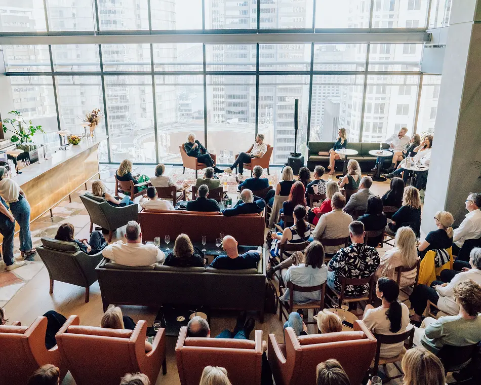 An audience sits in armchairs attending a talk inside a spacious, high-rise room featuring large floor-to-ceiling windows overlooking a city skyline.