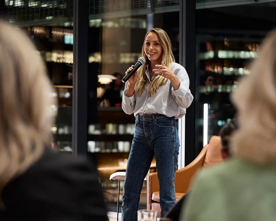 A smiling woman wearing a striped shirt and jeans speaks into a microphone while gesturing, standing before an audience inside an evening event space with large glass windows.