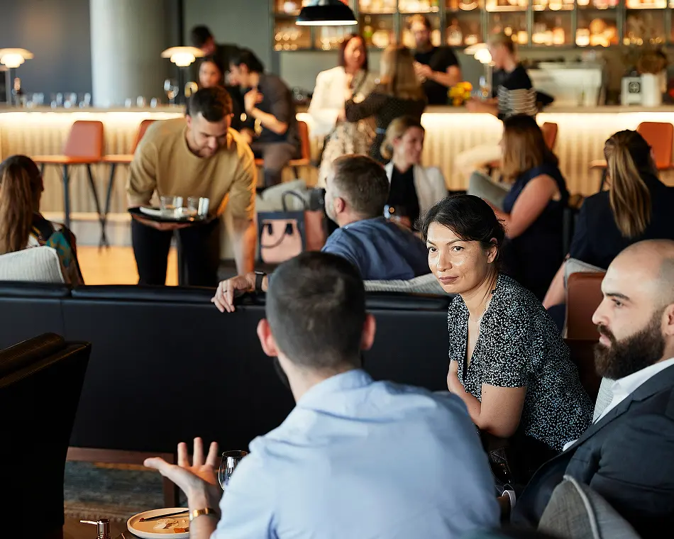 Patrons converse while sitting around couches in a stylish, dimly lit lounge as a waiter carries a tray of drinks near an illuminated bar in the background.