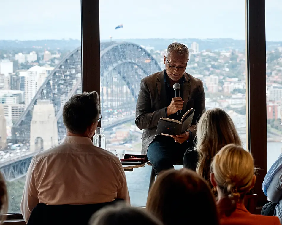 A man in glasses and a blazer speaks into a microphone while reading a book to an audience, seated in front of large windows overlooking the Sydney Harbour Bridge.