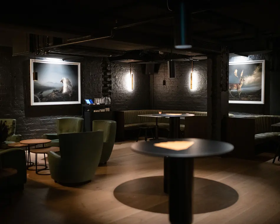 Green armchairs and black high-top tables furnish a dimly lit lounge with light wood floors, dark brick walls, subtle warm lighting, and framed animal artwork on the walls.