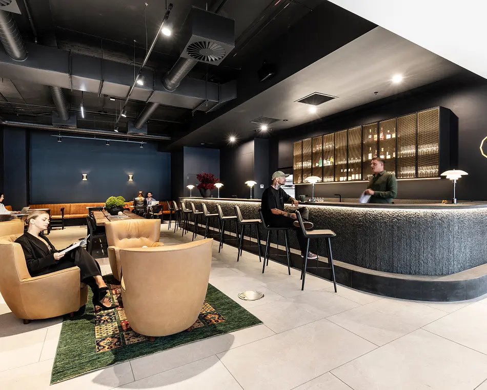 A woman reads a magazine in a leather armchair as a bartender converses with a patron at a curved dark bar inside a modern lounge with exposed ceiling ducts.