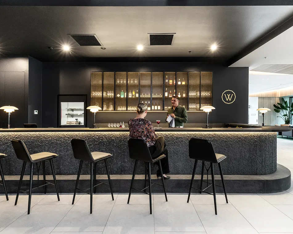 A bartender stands behind a dark, textured bar counter, attending to a customer seated on a barstool inside an elegant, dimly lit cocktail lounge. Text in the image includes: "W" and "Team only".