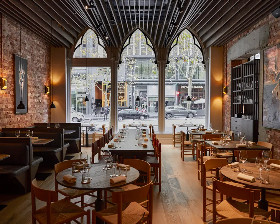 Elegantly set wooden dining tables and chairs fill a restaurant featuring exposed brick walls, slatted vaulted ceilings, and large gothic-arched windows overlooking a street displaying text reading "COFFEE", "FREYA", and "ROYAL STACKS".