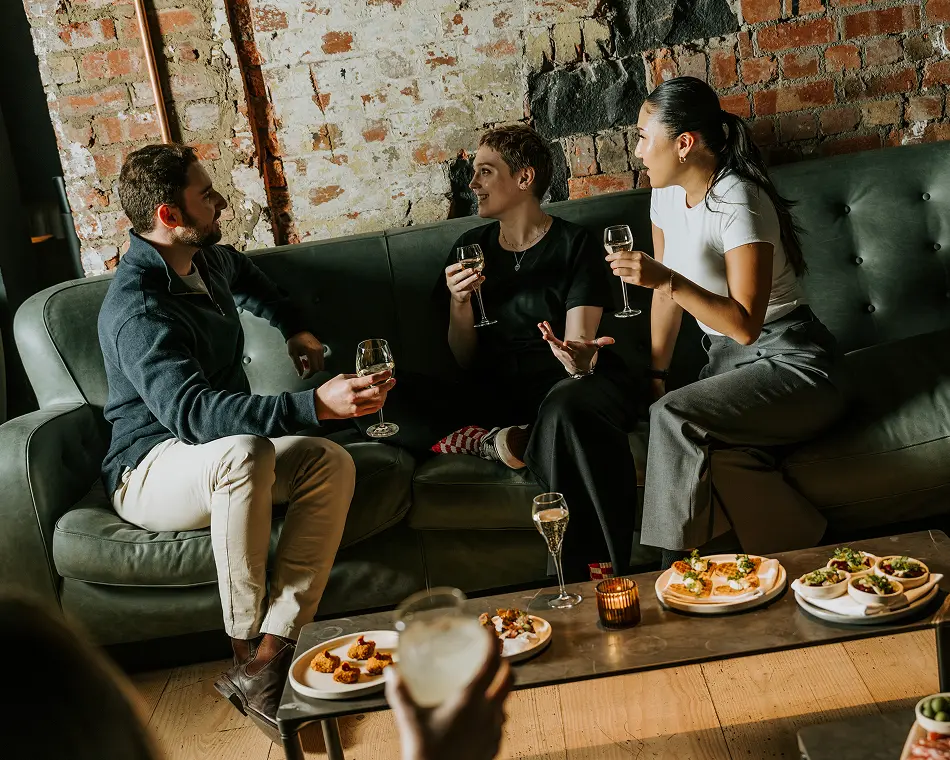 Three people sit on a green tufted leather sofa, drinking wine and talking together in front of an exposed brick wall, while a coffee table displays various small food plates.