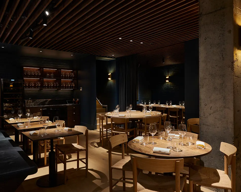 Set wooden dining tables and woven chairs fill a dimly lit restaurant dining space, surrounded by dark walls, warm accent lights, slatted wooden ceilings, and a concrete pillar.