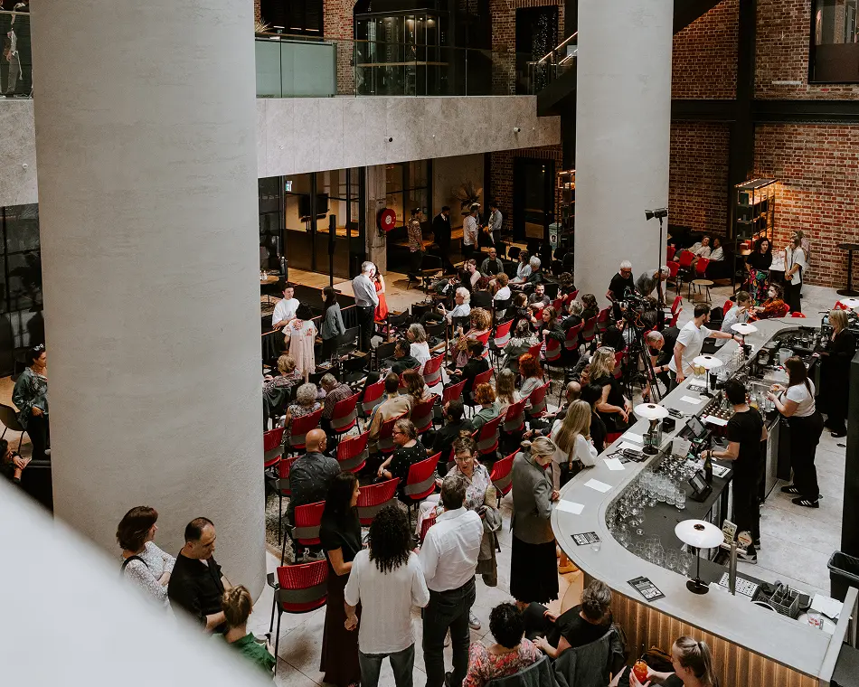 A large audience gathers in red chairs and around a curved bar to attend an event inside a spacious, modern atrium featuring prominent white columns and exposed brick walls.