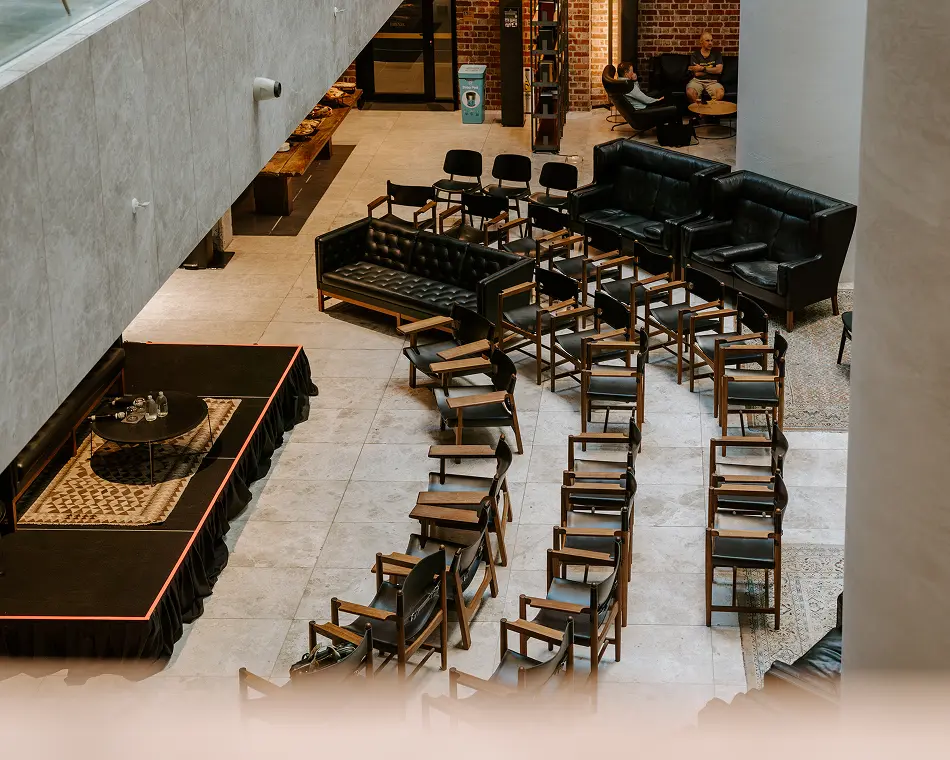 Rows of wooden and black leather chairs are arranged facing a small raised stage inside a spacious, modern atrium with tiled floors, brick walls, and leather sofas.