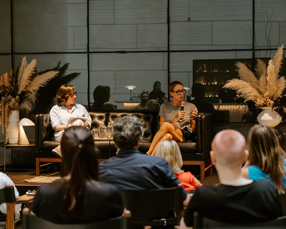 Two women sit on a black leather couch during a panel presentation, with one speaking into a microphone facing an audience in a modern indoor event space.