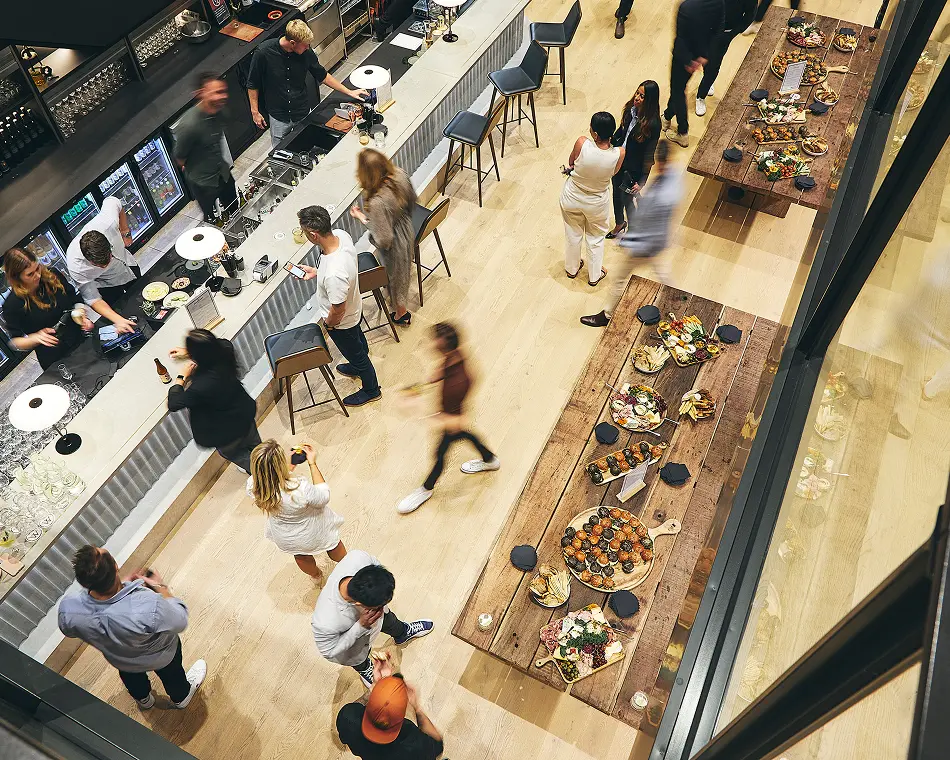People socialize, order drinks at a long bar, and enjoy food platters on rustic wooden tables inside a modern, brightly lit event space viewed from a high angle.