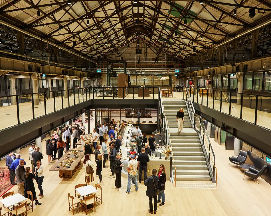 A crowd of people mingles and walks down stairs inside a large, multi-level industrial hall featuring exposed steel roof trusses and light wood floors.