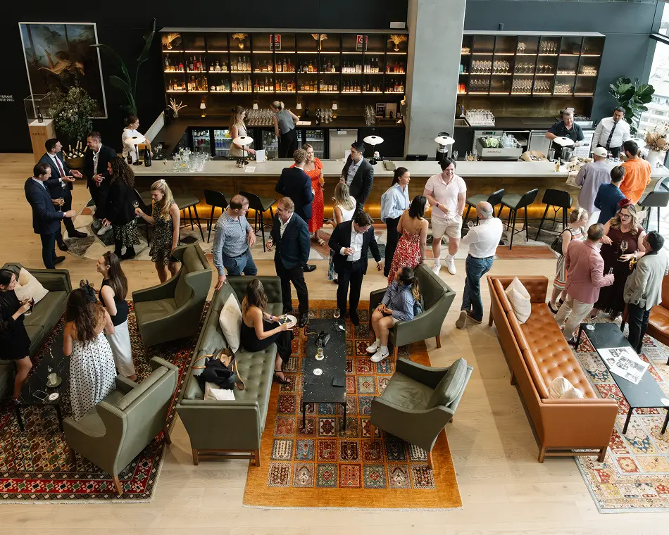 Well-dressed guests mingle, chat, and enjoy drinks in a spacious, modern lounge filled with plush leather sofas, armchairs, colorful area rugs, and a long, fully stocked bar background.