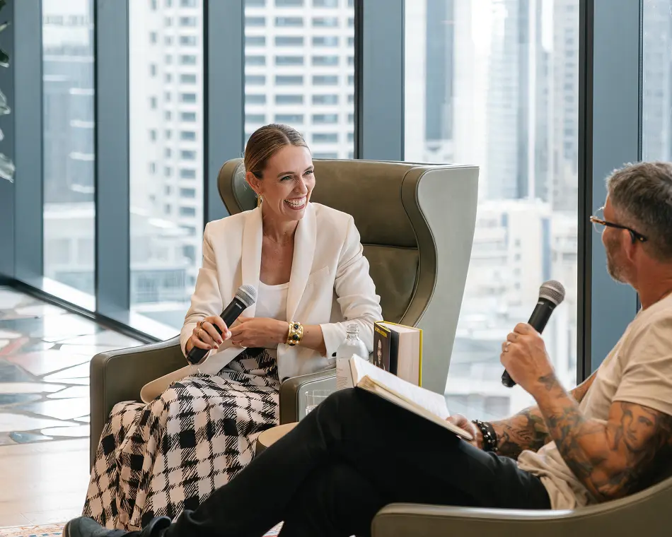 A smiling woman wearing a white blazer and a tattooed man sit in armchairs, holding microphones and conversing in a modern room with floor-to-ceiling windows overlooking a city skyline.