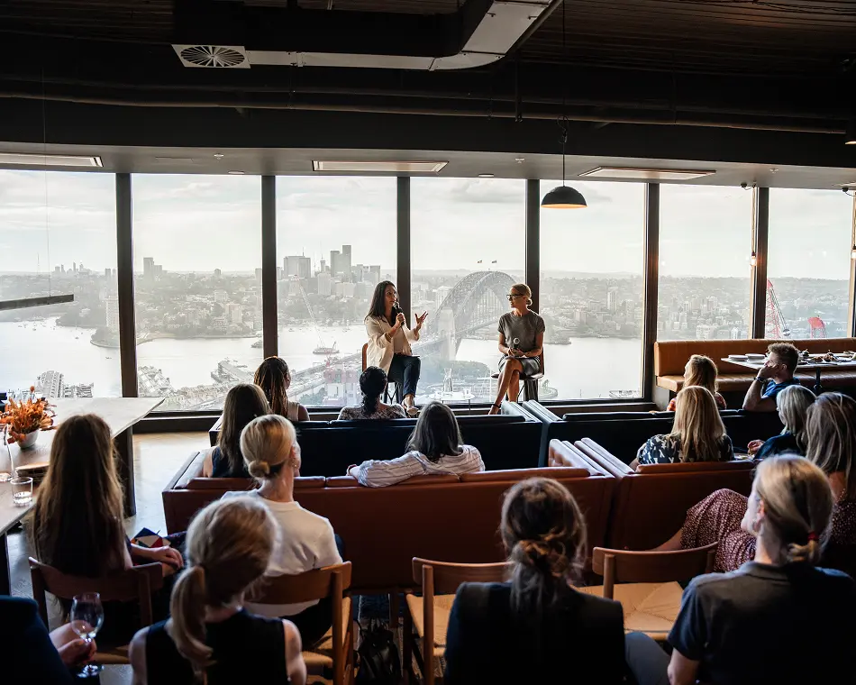 Two women speak into microphones during a presentation to a seated audience inside a high-rise event space featuring large floor-to-ceiling windows that overlook a sprawling harbor city skyline.