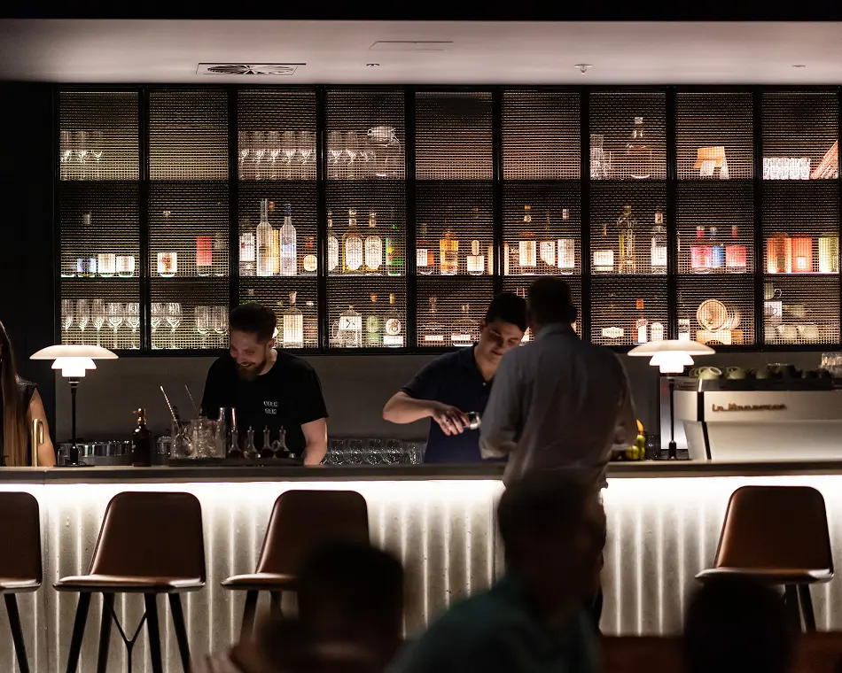Two bartenders prepare drinks behind a brightly lit bar counter in a stylish lounge with empty barstools and a large, grid-patterned wall shelf filled with illuminated bottles. Text on the espresso machine reads: "La Marzocco".