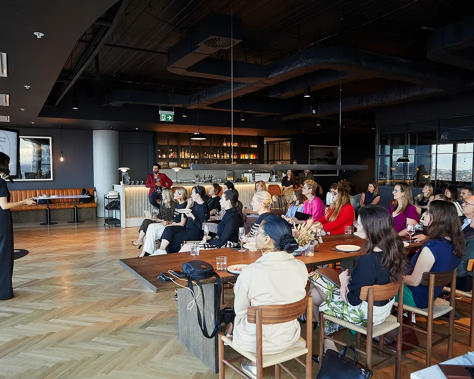 An audience of seated professionals listens to a presenter at a long wooden table inside a modern venue with herringbone wood floors and a bar in the background.