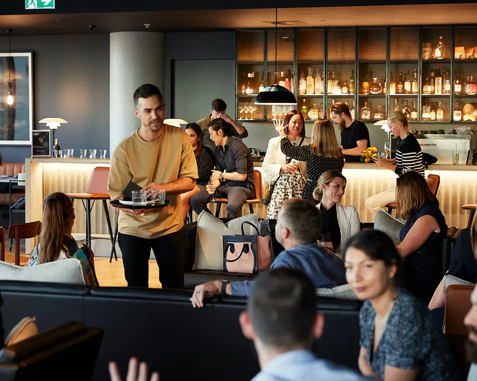 A waiter in a beige shirt carries a tray of drinks through a crowded, modern lounge filled with seated guests socializing near a lit bar.