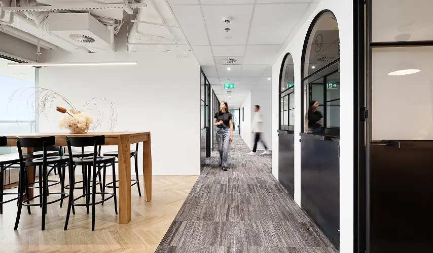 A woman in a black shirt walks down a carpeted hallway past a high wooden table with barstools and arched glass partitions inside a bright, modern office space.