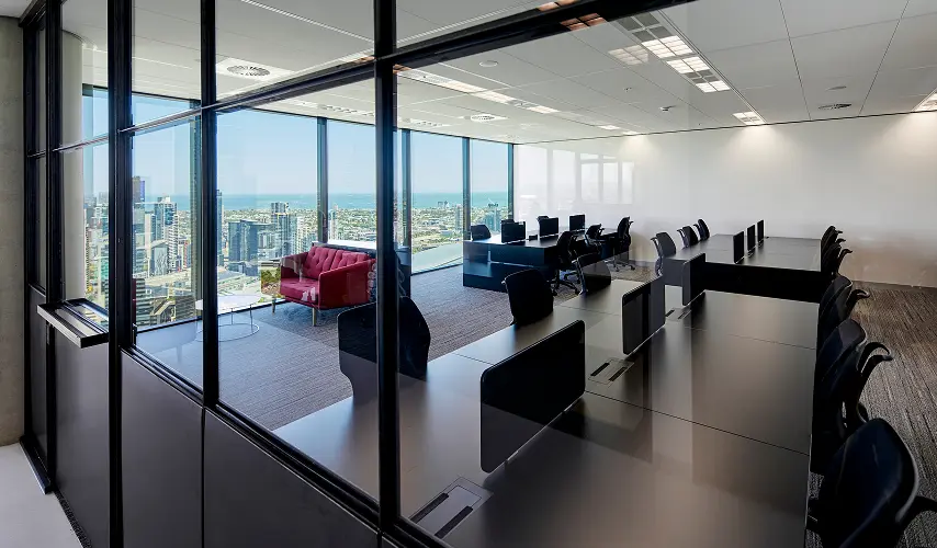 Sleek dark office desks lined with black chairs sit inside a glass-partitioned high-rise workspace overlooking a panoramic city skyline and ocean view through floor-to-ceiling windows.