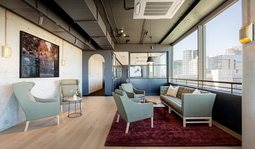 Sage green armchairs and a tufted sofa sit on a red area rug within a modern office lounge featuring concrete walls, exposed ceiling ducts, and large city-view windows.