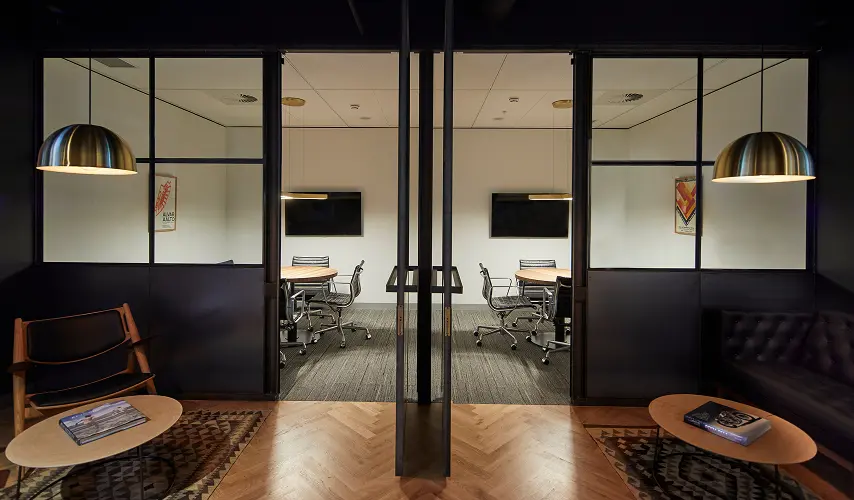 Two glass-partitioned meeting rooms containing round tables and chairs sit side-by-side behind symmetrical lounge areas with coffee tables and pendant lights. A framed wall poster reads "ALVAR AALTO".