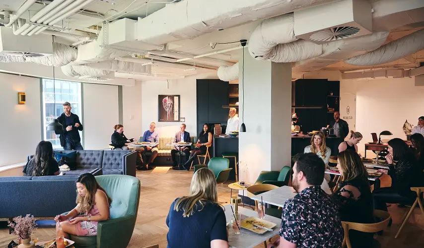 People sit, dine, and converse at various tables and armchairs inside a modern open-plan lounge featuring exposed ceiling ductwork, wooden floors, and a wall sign reading "WORK CLUB".