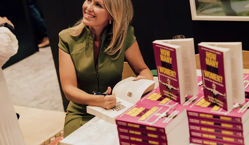 A blonde woman in an olive green dress smiles while signing a copy of her book at a marble table, surrounded by stacks of the publication at a book signing event. The books display the text: "INTERNATIONAL BESTSELLER UPDATED", "HOW MANY MORE WOMEN?", "How the law silences women", and "Jennifer Robinson & Keina Yoshida".