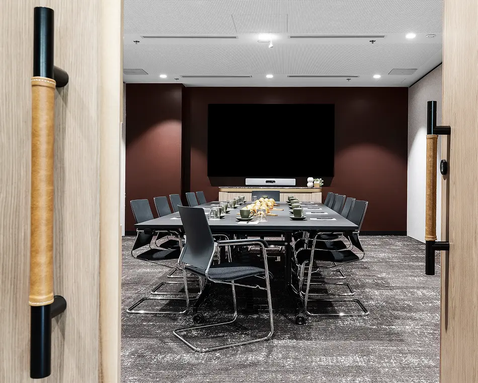 A long conference table set with refreshments and surrounded by chairs sits inside a modern boardroom with a large wall-mounted screen, viewed between open wooden doors.