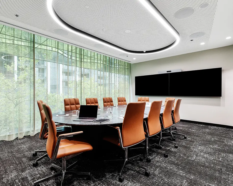 An oval conference table surrounded by tan leather chairs holds a laptop and water pitcher in a modern boardroom featuring an oval ceiling light and large sheer-curtained windows.