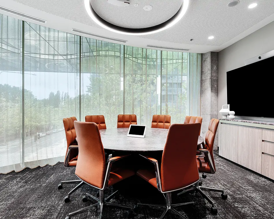 A round marble conference table, surrounded by brown leather office chairs, sits centered in a modern meeting room featuring floor-to-ceiling glass windows, dark carpet, and a circular ceiling light.