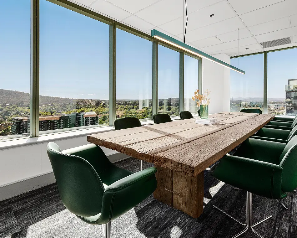 A long rustic wooden conference table sits surrounded by dark green swivel armchairs in a modern office featuring floor-to-ceiling windows that offer expansive views of a city and mountains.
