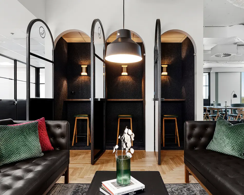 Three arched, dark-paneled phone booths featuring wooden stools stand behind a lounge area with black leather sofas and a coffee table with a green vase. Text on the book on the table reads: "PRINCIPLES OF STYLE", "RICHARD AINSWORTH".