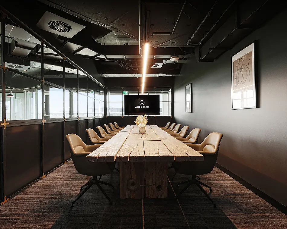 A long rustic wooden conference table surrounded by beige swivel chairs extends through a modern boardroom with dark walls, exposed ceiling pipes, and glass partitions. Text on the TV screen reads: "W WORK CLUB". Text on the framed art reads: "FEAST".