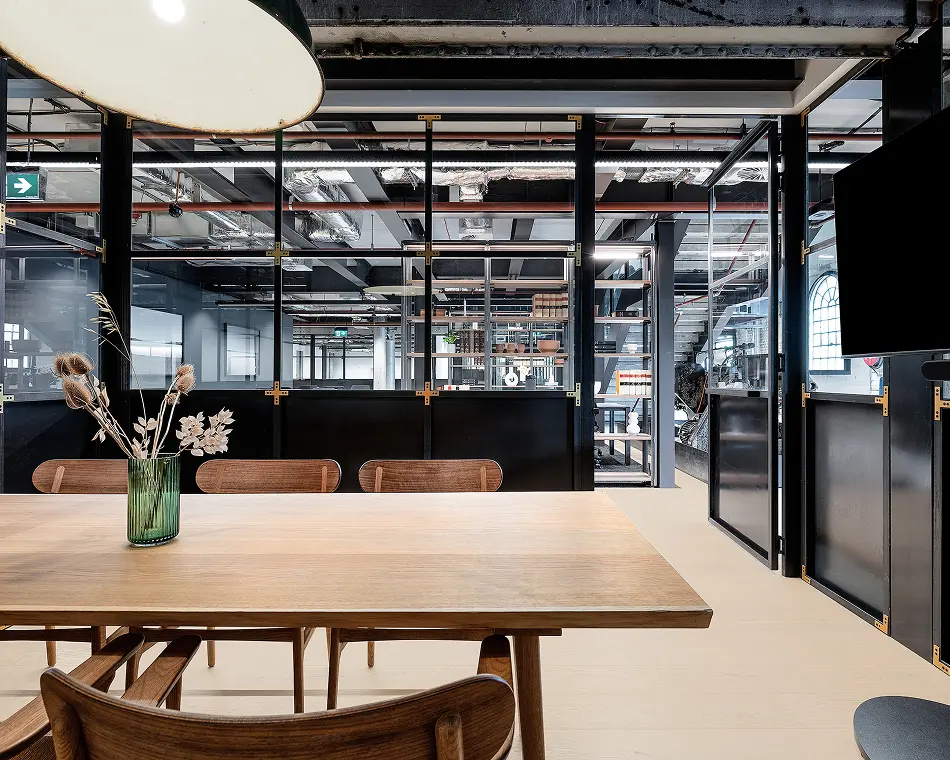A light wooden conference table surrounded by chairs sits in a modern office, bounded by black-framed glass partition walls that reveal an industrial space with exposed ceiling pipes.