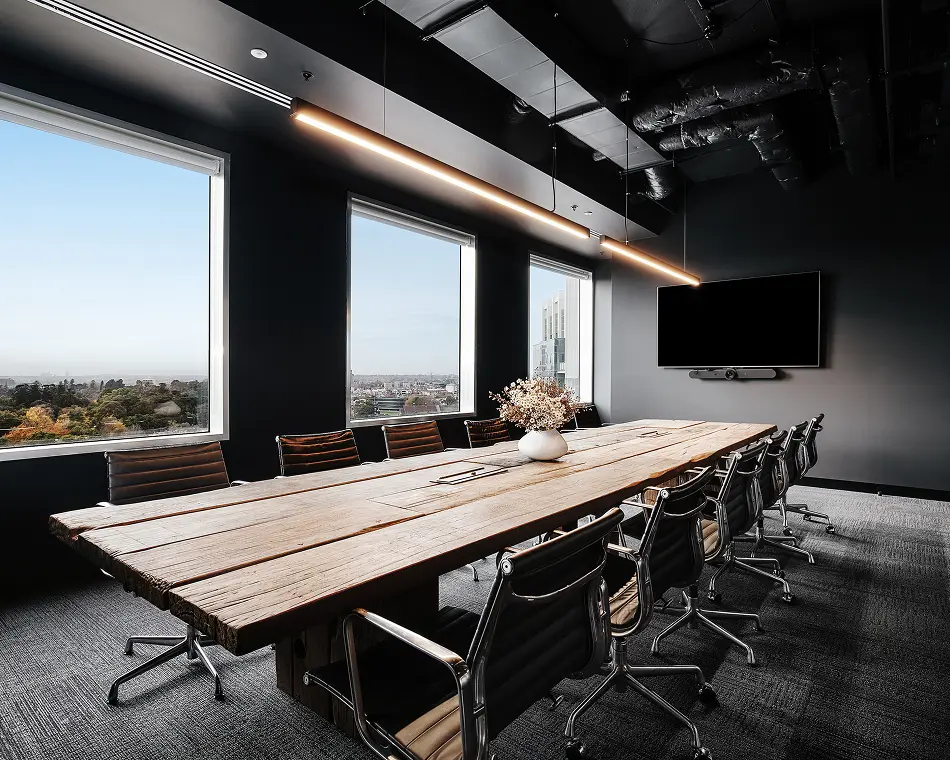 A long wooden conference table surrounded by black leather chairs extends through a modern boardroom featuring dark walls, linear pendant lighting, and large windows overlooking an expansive city view.