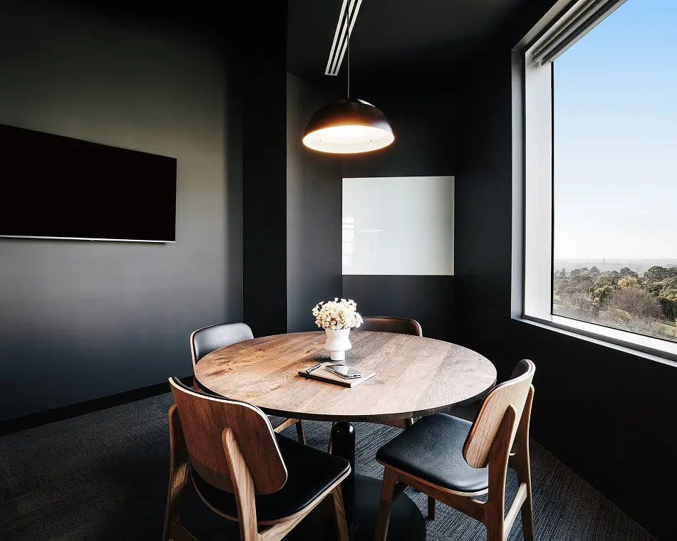 A round wooden table surrounded by four chairs sits illuminated under a pendant light inside a dark, modern conference room featuring a large window with an elevated city view.