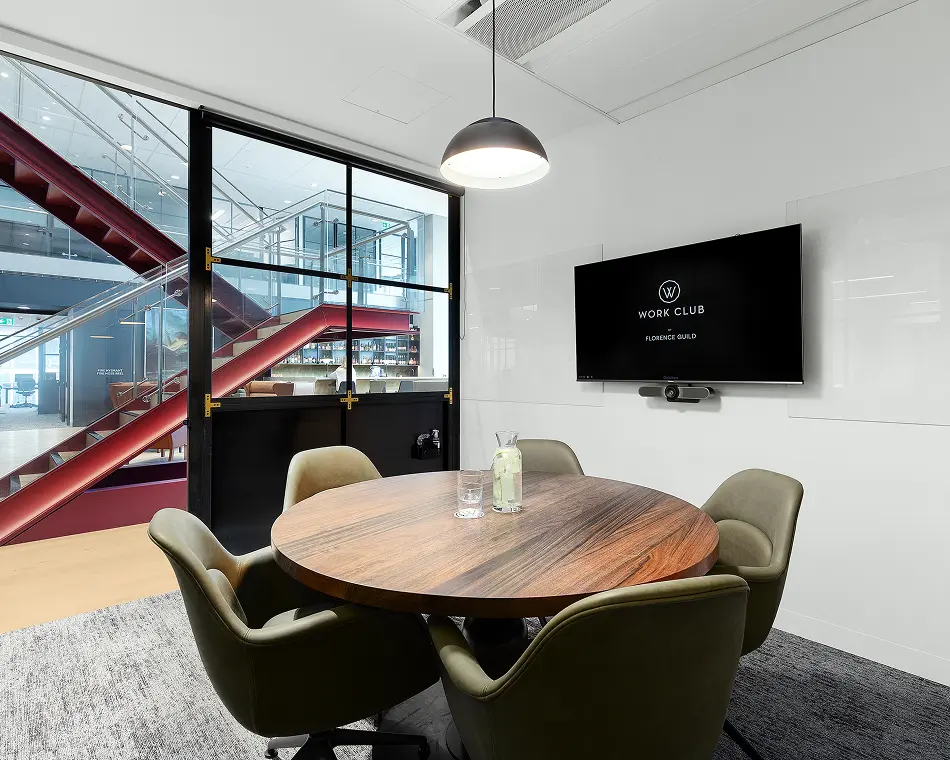 A round wooden meeting table surrounded by olive-green chairs stands under a pendant light in a modern office room equipped with a wall-mounted monitor displaying "W WORK CLUB BY FLORENCE GUILD".